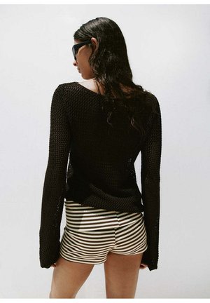 Femme aux longs cheveux foncés portant un pull en maille noir, des lunettes de soleil noires et un short beige et noir à rayures, de dos sur fond blanc.