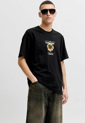 Tânăr purtând ochelari de soare negri, un tricou negru cu un grafic de floarea-soarelui și text, și blugi închiși la culoare, decolorați, cu o mână în buzunar.