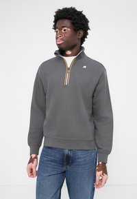 Sweat-shirt gris avec un col montant et une fermeture éclair quart. Caractéristiques de rayures contrastantes jaunes et bleues sur la fermeture éclair et un petit logo sur la poitrine.