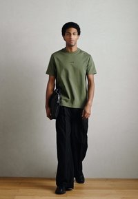 Olivzaļš īsroku T-krekls ar apaļu kaklu, kopa ar vaļīgām melnām biksēm un spīdīgiem melniem apaviem. Zem rokas tiek turēta melna soma.