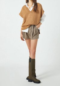 Chaleco de punto marrón sobre una camisa blanca de cuello, acompañado de pantalones cortos de cuadros marrones y botas de lluvia olivas con acentos negros y suelas texturizadas.