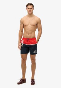 Shorts de baño en rojo y azul marino con cintura ajustable, que presentan detalles en blanco y el logotipo de texto "SUPER DRY CO." en la pierna izquierda.