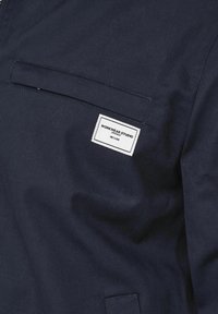 Giacca da lavoro blu navy realizzata in tessuto robusto, con una tasca sul petto dotata di un'etichetta bianca con la scritta "WORKWEAR STUDIO". Dettagli di cuciture semplici.