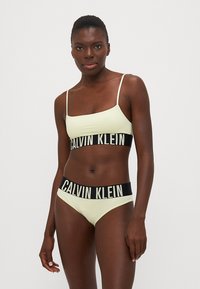 Lysegrønn Calvin Klein bikini med sort logobånd. Toppen har tynne stropper og et cropped design, mens bunnen er en lavt livsbrief.