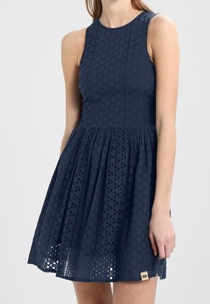 Granatowa sukienka na ramiączkach z ażurowym wzorem, z dopasowanym gorsetem i rozkloszowaną spódnicą, noszona przez stojącą osobę z ramieniem swobodnie opuszczonym wzdłuż ciała.