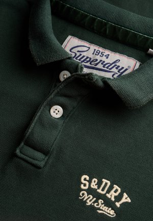 Polo shirt verde scuro con tessuto testurizzato, due bottoni bianchi e ricamo "S&Dry" in crema. L'etichetta riporta "Superdry 1954."