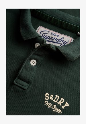 Polo shirt verde scuro con tessuto testurizzato, due bottoni bianchi e ricamo "S&Dry" in crema. L'etichetta riporta "Superdry 1954."
