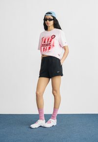 Žena odjevena u ružičastu Nike majicu s grafičkim printom, crne Nike kratke hlače, ružičaste čarape, bijele tenisice, plavu kapu naopačke i crne sunčane naočale stoji na plavom podu.