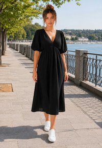 Femme avec les cheveux en chignon, vêtue d'une robe midi noire et de baskets blanches, marchant le long d'une promenade au bord de la rivière avec des arbres et une rampe en métal.