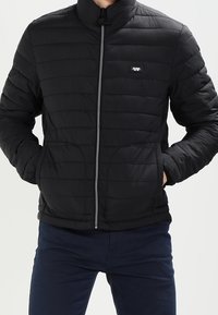 Černá péřová bunda s prošívaným designem, vysokým límcem a zipovým zapínáním. Obsahuje přední kapsy a malé logo na hrudi.