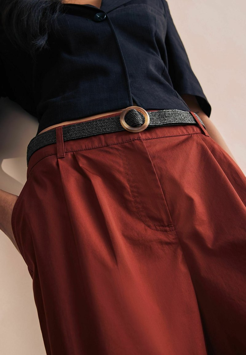 Femme portant un pantalon taille haute de couleur rouille avec une ceinture tissée noire avec une grande boucle ronde et un haut court noir à boutons.