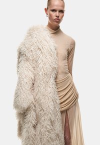 Beige nep-bontjas met lange, harige textuur, gedragen over een aansluitende nude mesh top. De rok heeft gedrapeerd materiaal en transparante elementen.