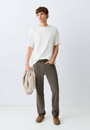 Jeune homme portant un t-shirt blanc et un pantalon marron tient une veste beige dans une main, l'autre main dans sa poche, debout sur un sol blanc.