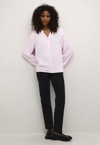 Blouse rose clair à manches longues avec un col en V et une patte de boutonnage à trois boutons, associée à un pantalon noir droit et des mocassins noirs.