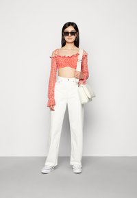 Top curto floral vermelho com bordas franzidas, combinado com calças brancas de perna larga e ténis brancos. O modelo segura uma mala de ombro creme.