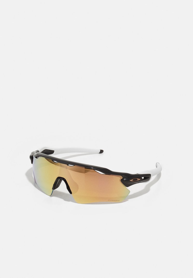 Lunettes de soleil avec une monture noir et blanc, de grandes lentilles réfléchissantes aux nuances dorées et roses, un design aérodynamique et un bord extérieur élargi.