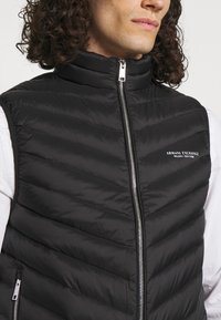 Svart vadderad väst med hög krage, med chevron-quilting, en hel frontdragkedja och en logotyp på vänster bröstområde.