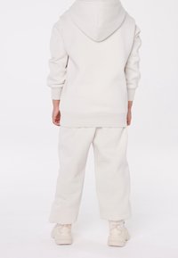 Ett barn står vänd bort med en off-white huva sweatshirt, matchande lösa byxor och beige sneakers mot en vit bakgrund.