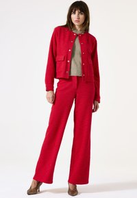 Veste texturée rouge avec un pantalon texturé rouge assorti. La veste présente des boutons-pression et des poches appliquées, associée à un pull gris en dessous.