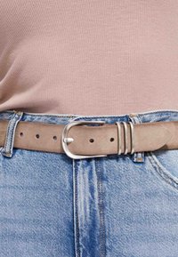 Beiger Wildledergürtel mit silberner Schnalle, der über hellblauen Jeans und in ein geripptes, hellrosa Oberteil gesteckt ist.