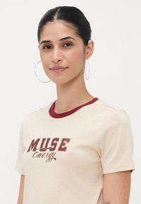 Cream cotton t-shirt with maroon ribbed collar and "MUSE Energy" graphic in bold lettering, short sleeves, and soft texture.
