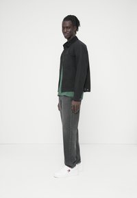 Νεαρός άνδρας σε πλάγια στάση, φοράει μαύρο μπουφάν, πράσινο πουκάμισο, γκρι τζιν και λευκά αθλητικά παπούτσια σε λευκό φόντο.