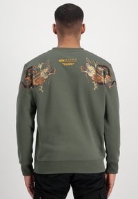 Felpa verde oliva con maniche lunghe, caratterizzata da un intricato design grafico sulla schiena e un orlo a costine. Marchio posizionato nella parte alta della schiena.