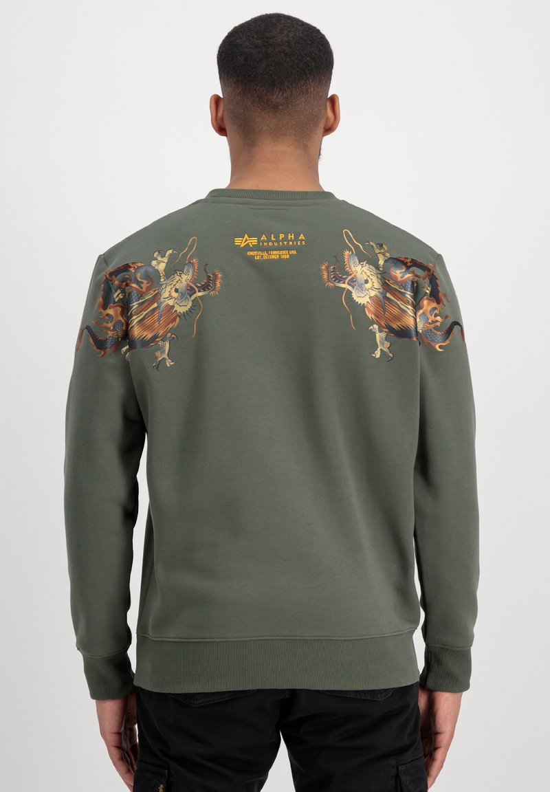 Felpa verde oliva con maniche lunghe, caratterizzata da un intricato design grafico sulla schiena e un orlo a costine. Marchio posizionato nella parte alta della schiena.