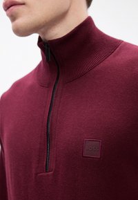 Muškarac nosi tamnocrveni zip-up pulover s visokim rebrastim ovratnikom i "BOSS" logotipom na prsima.