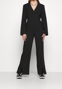 Mono negro con mangas largas, escote en forma de V, corte entallado y aberturas en los tobillos. Combinado con zapatillas negras y blancas.