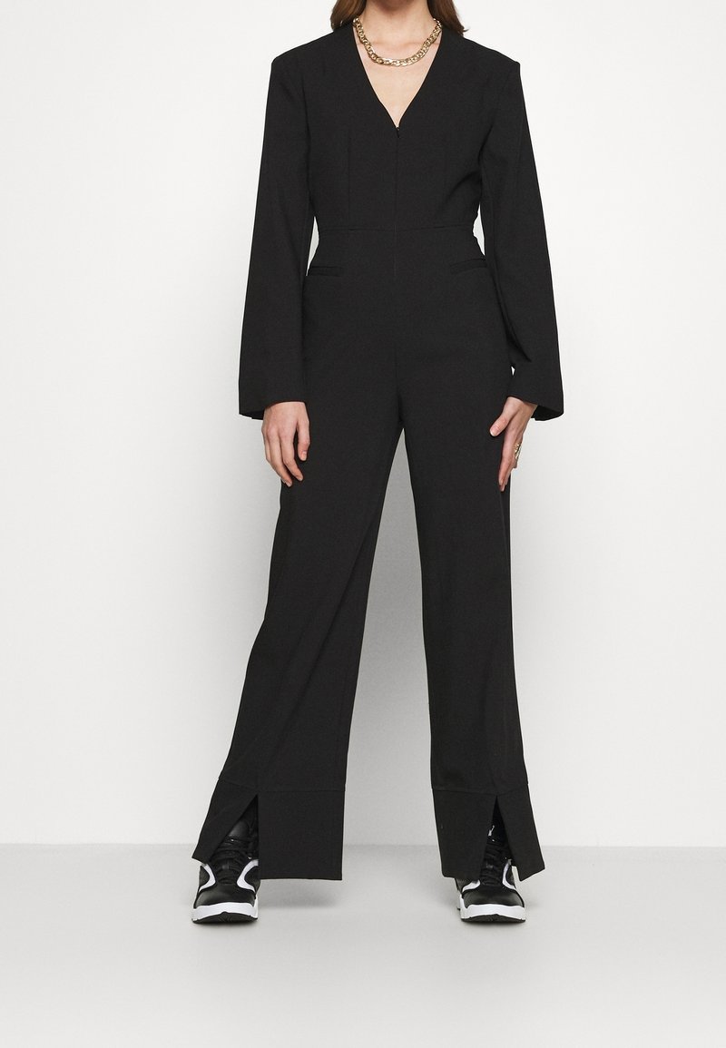Mono negro con mangas largas, escote en forma de V, corte entallado y aberturas en los tobillos. Combinado con zapatillas negras y blancas.
