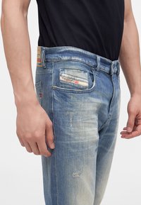 Homme portant un jean en denim bleu délavé avec une étiquette sur la petite poche avant et une chemise noire rentrée.