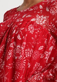 Tissu rouge avec un motif floral blanc ; coupe décontractée avec des détails plissés au niveau de la poitrine. Doté d'un col rond et d'une texture légère.