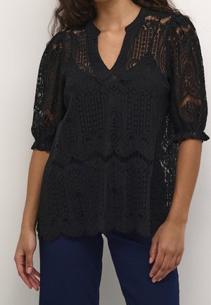 Femme portant un chemisier en dentelle noire avec des manches bouffantes et des bords festonnés, associée à un pantalon bleu foncé, debout devant un fond uni.