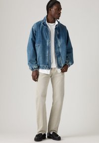 Man with braided hair wearing a blue denim jacket, white shirt, light beige pants, and black loafers standing against a plain background.