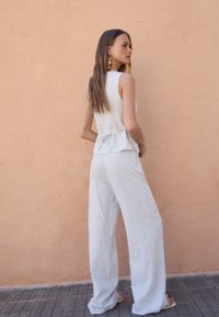 Lys grå, struktureret tanktop med peplum-design, parret med bredbenede, mønstrede bukser. Sat op imod en ferskenfarvet væg.