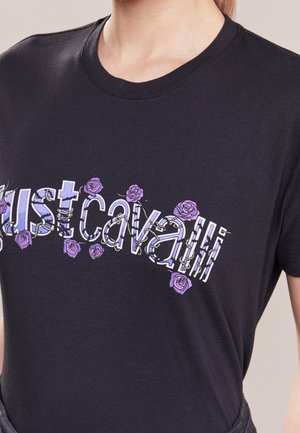 Chemise noire avec texte graphique floral blanc et violet disant "just cavalli", portée par une personne montrée du cou à la taille.