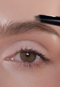Primo piano di un sopracciglio mentre viene stilizzato con un applicatore nero. Presenta peli scuri sul sopracciglio, pelle con un leggero trucco e un effetto sfocato sugli occhi.