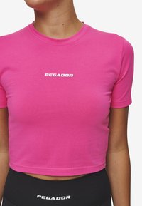 Ružičasta kratka majica s okruglim izrezom i kratkim rukavima. Na prednjoj strani nalazi se bijeli logo "PEGADOR". Glatka, rastezljiva tkanina.