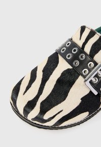 Musta ja kerma zebra-kuosiset klomppikengät, joissa on musta hihna hopeisella kiinnityksellä ja pyöreillä rei'illä, valmistettu teksturoidusta materiaalista.
