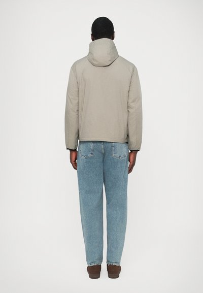 Helehall, tekstuuriga jope kapuutsiga, mis on kombineeritud lahtiste helesiniste teksade ja pruunide jalanõudega; vaadatuna tagantpoolt valgel taustal.