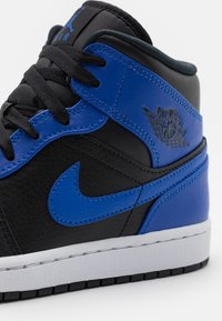 Primer plano de unas zapatillas Nike Air Jordan en negro y azul, mostrando los cordones, las costuras, los logotipos y la entresuela blanca sobre un fondo blanco.