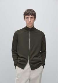 Dark olive green zip-up sweater with a high collar, featuring ribbed texture at shoulders and cuffs, worn over light beige pants.