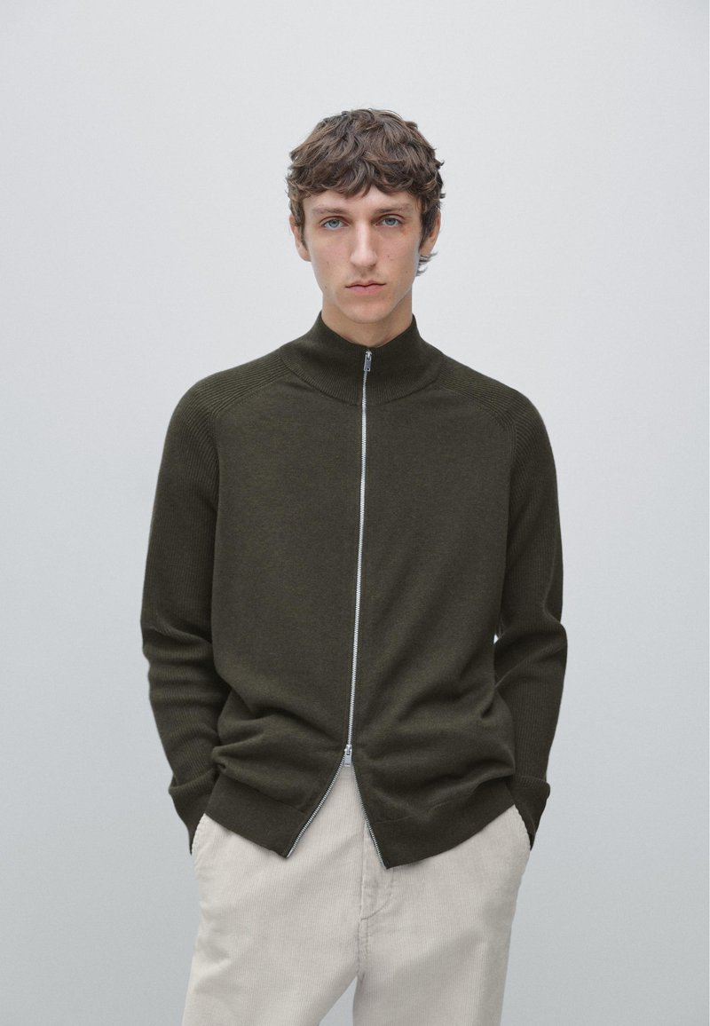 Dark olive green zip-up sweater with a high collar, featuring ribbed texture at shoulders and cuffs, worn over light beige pants.