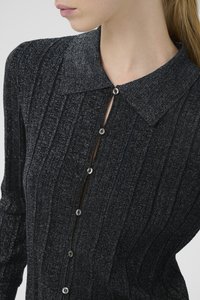 Közeli kép egy nőről, aki fekete textúrával rendelkező, gombos felsőt visel hegyes gallérral és ezüst gombokkal, látható a nyaka és az arca egy része.