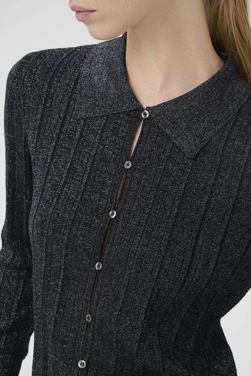 Nahaufnahme einer Frau, die ein schwarzes, strukturiertes Hemd mit Knöpfen und einem spitzen Kragen sowie silbernen Knöpfen trägt. Ihr Hals und ein Teil des Gesichts sind sichtbar.
