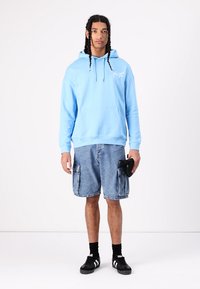 Felpa con cappuccio azzurro chiaro con tasca frontale e lacci, abbinata a pantaloni cargo in denim blu con tasche laterali e sneakers nere.