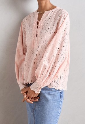 Femme portant un chemisier en dentelle à œillets rose pâle avec des bords festonnés et des manches larges, assorti à un jean en denim bleu.