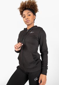 Zwarte ritshoodie van gladde stof, met een capuchon, voorzakken en een wit logo op de borst, gecombineerd met zwarte legging.