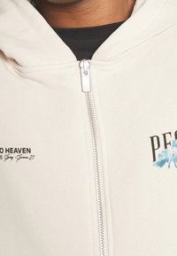 Krémová mikina s kapucí na plný zip, s černým potištěným textem a modrým grafickým akcentem. Povrch vypadá měkce a připomíná bavlnu.